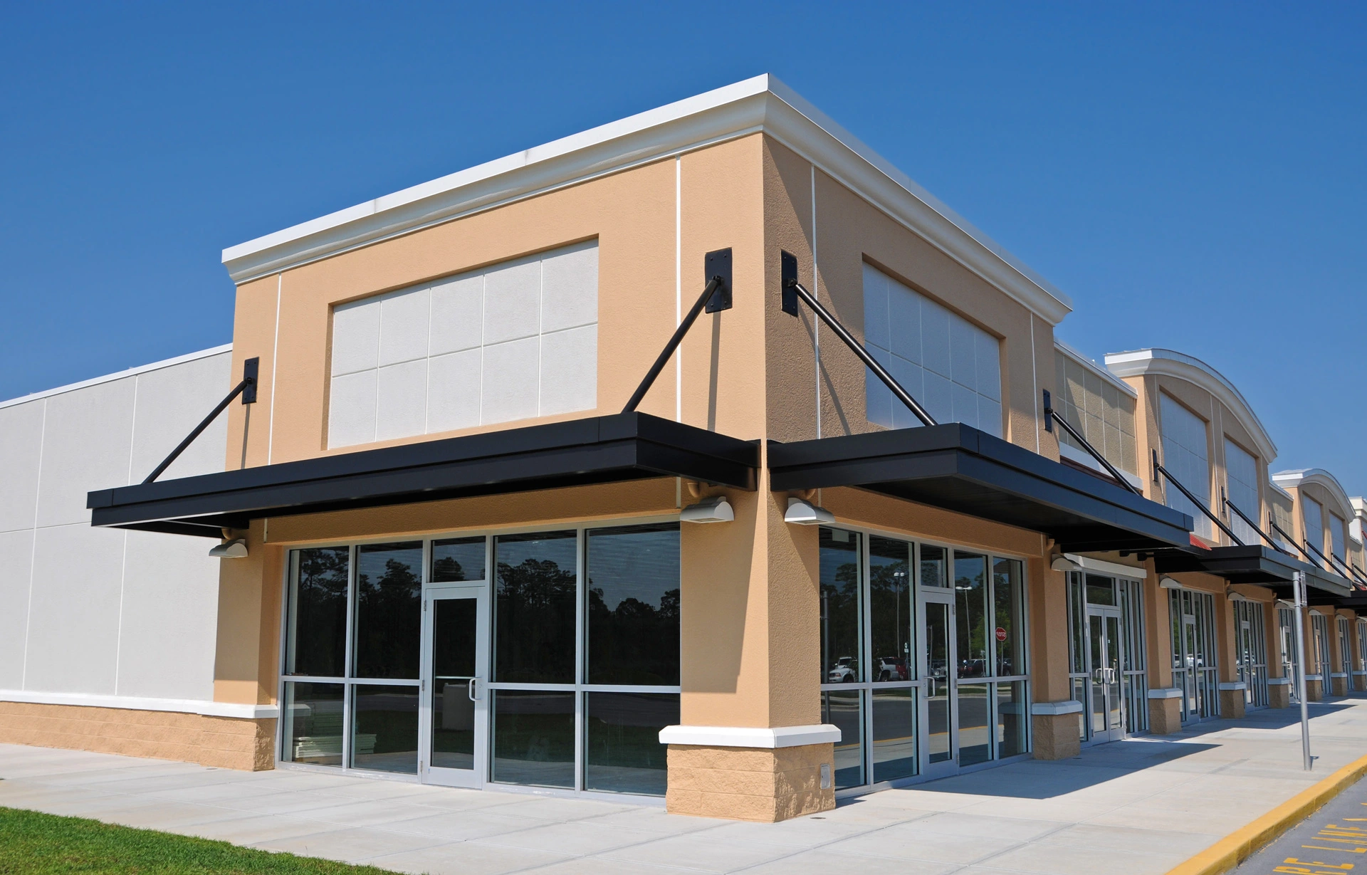 Exterior corner view of a vacant, modern commercial retail building featuring tan stucco walls, large glass windows, and black metal awnings.