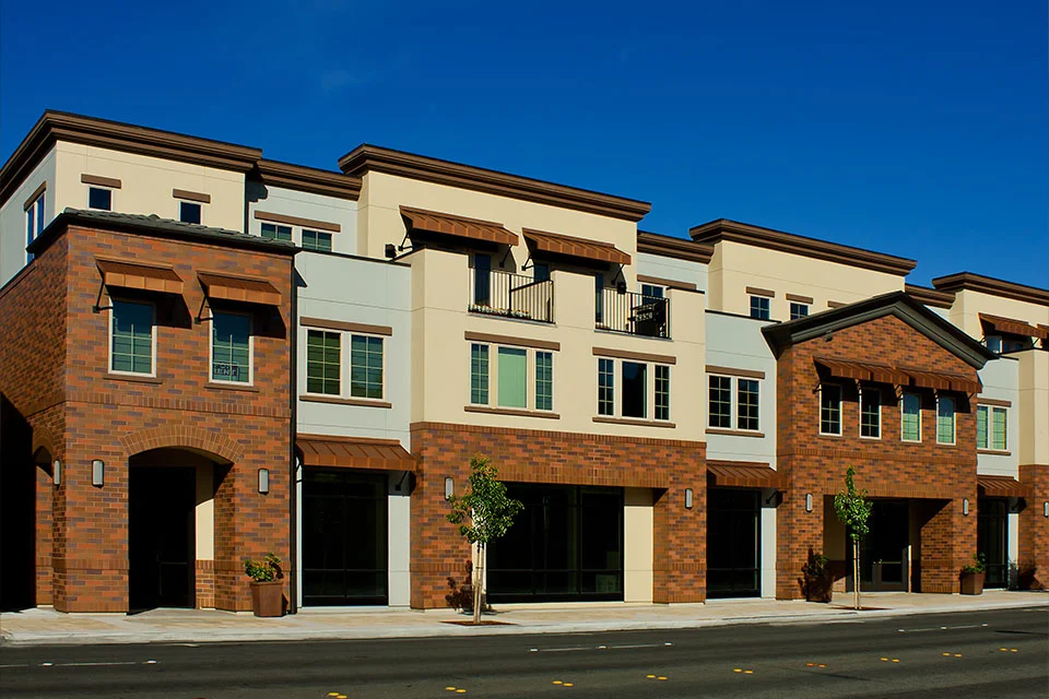 Exterior street view of a modern multi-story commercial and residential building featuring tan stucco, brown brick ground-floor facades, and metal window awnings on a sunny day.