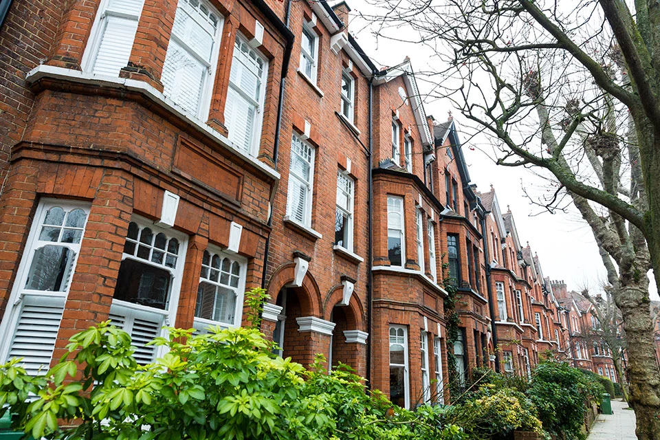 Exterior street view of a row of traditional red brick townhouses featuring white window trim and green landscaping in the foreground.