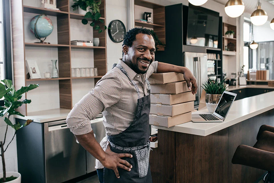 A smiling business owner wearing an apron leans against a stack of shipping boxes next to a laptop in a modern workspace.
