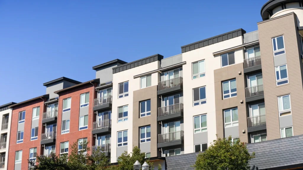 Low-angle exterior view of a modern multi-story apartment building featuring a mixed-color facade of red, cream, and tan with metal balconies under a blue sky.
