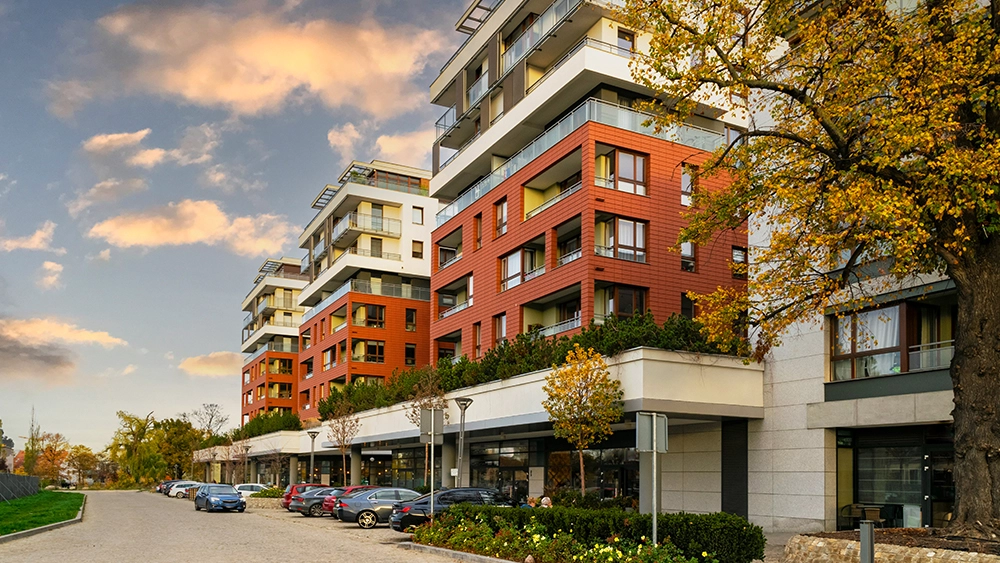 Exterior view of modern multi-story apartment buildings with reddish-orange facades, white balconies, and autumn trees alongside parked cars.